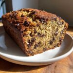 A loaf of banana zucchini bread with chocolate chips, sitting on a white plate.