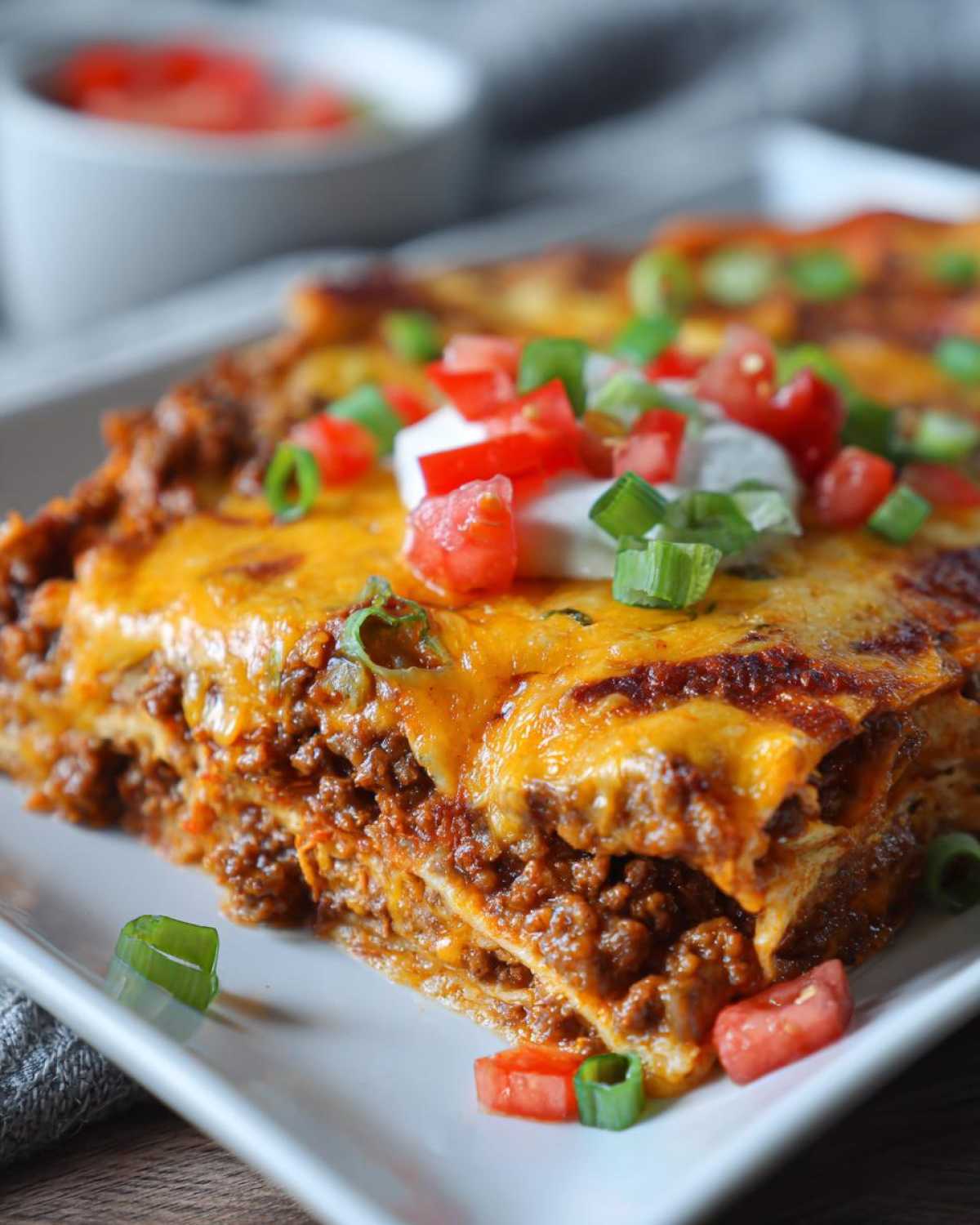 A slice of burrito bake layered with meat, cheese, and tortillas, topped with sour cream, tomatoes, and green onions.