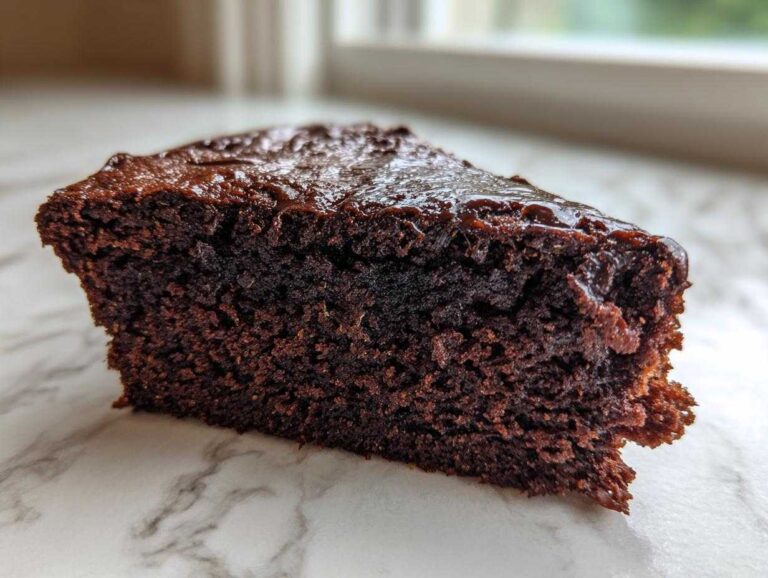 A slice of rich, fudgy chocolate zucchini cake with a glossy chocolate topping on a marble surface.