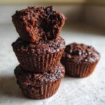 A stack of three chocolate zucchini muffins, one with a bite taken out, showing the moist interior.