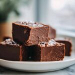A stack of homemade chocolate fudge recipe squares on a white plate, sprinkled with sea salt.