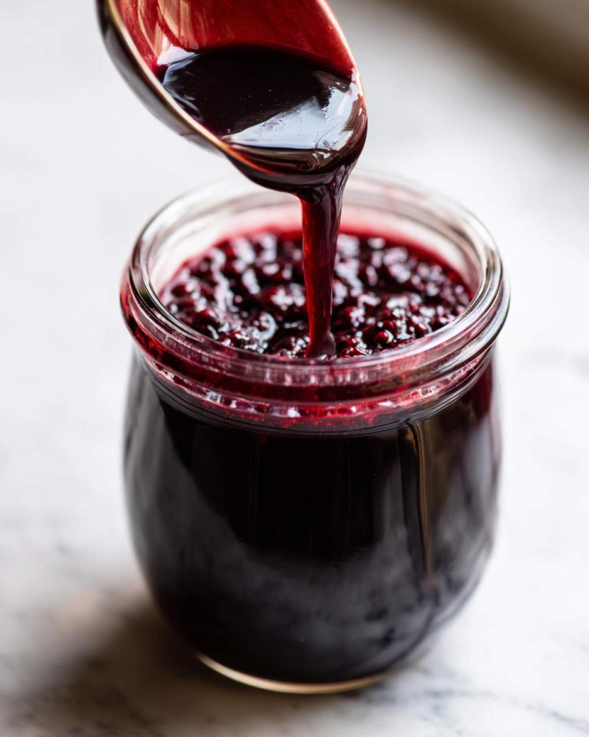 A spoon pouring dark purple elderberry syrup into a glass jar filled with elderberries.
