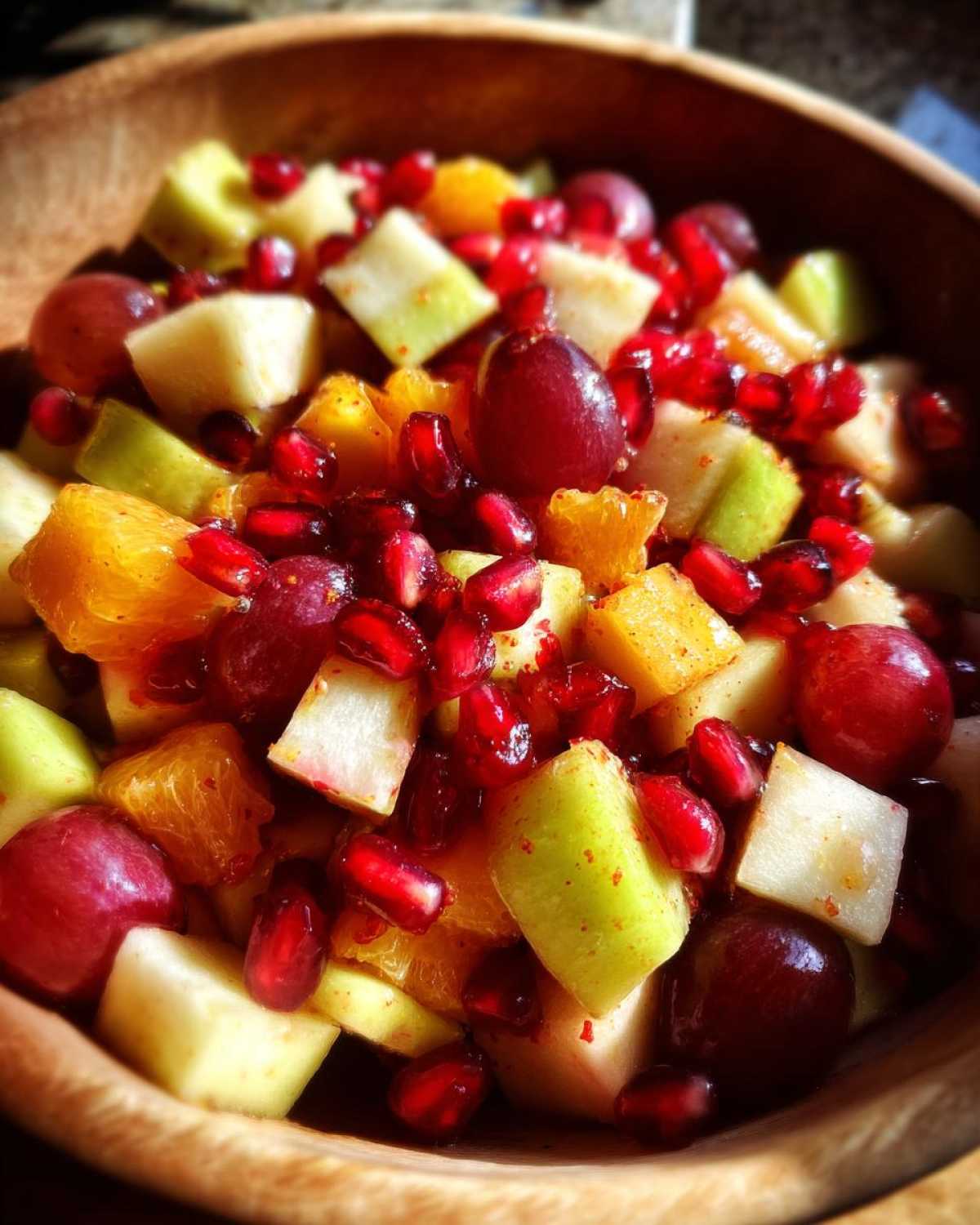 Close-up of a vibrant fall fruit salad in a wooden bowl, featuring apples, oranges, grapes, and pomegranate seeds.