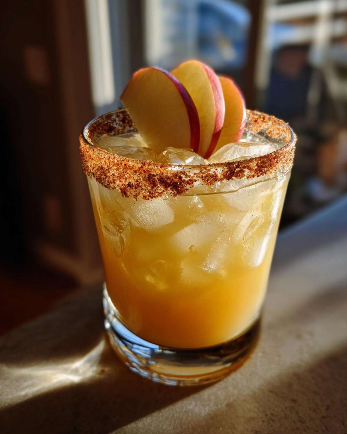 A vibrant fall margarita in a glass with a spiced rim and garnished with apple slices.