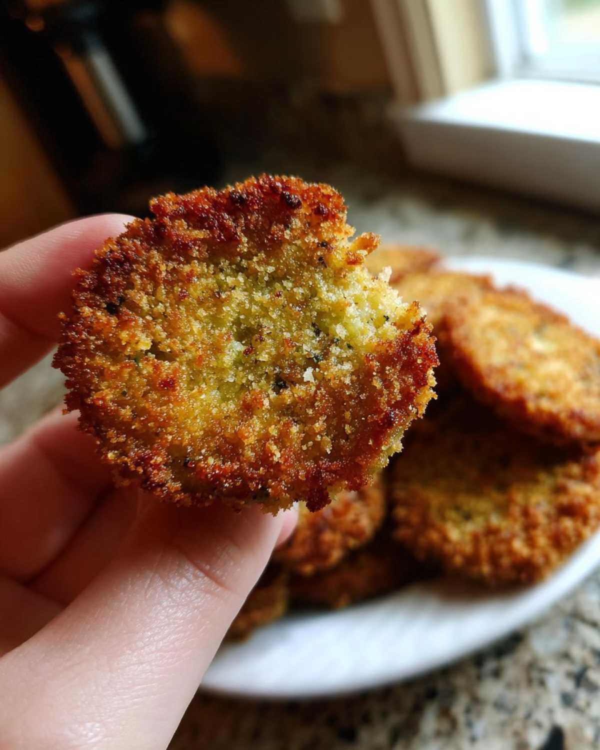 Close-up of a hand holding a crispy fried green tomato with a bite taken out, showcasing its texture.