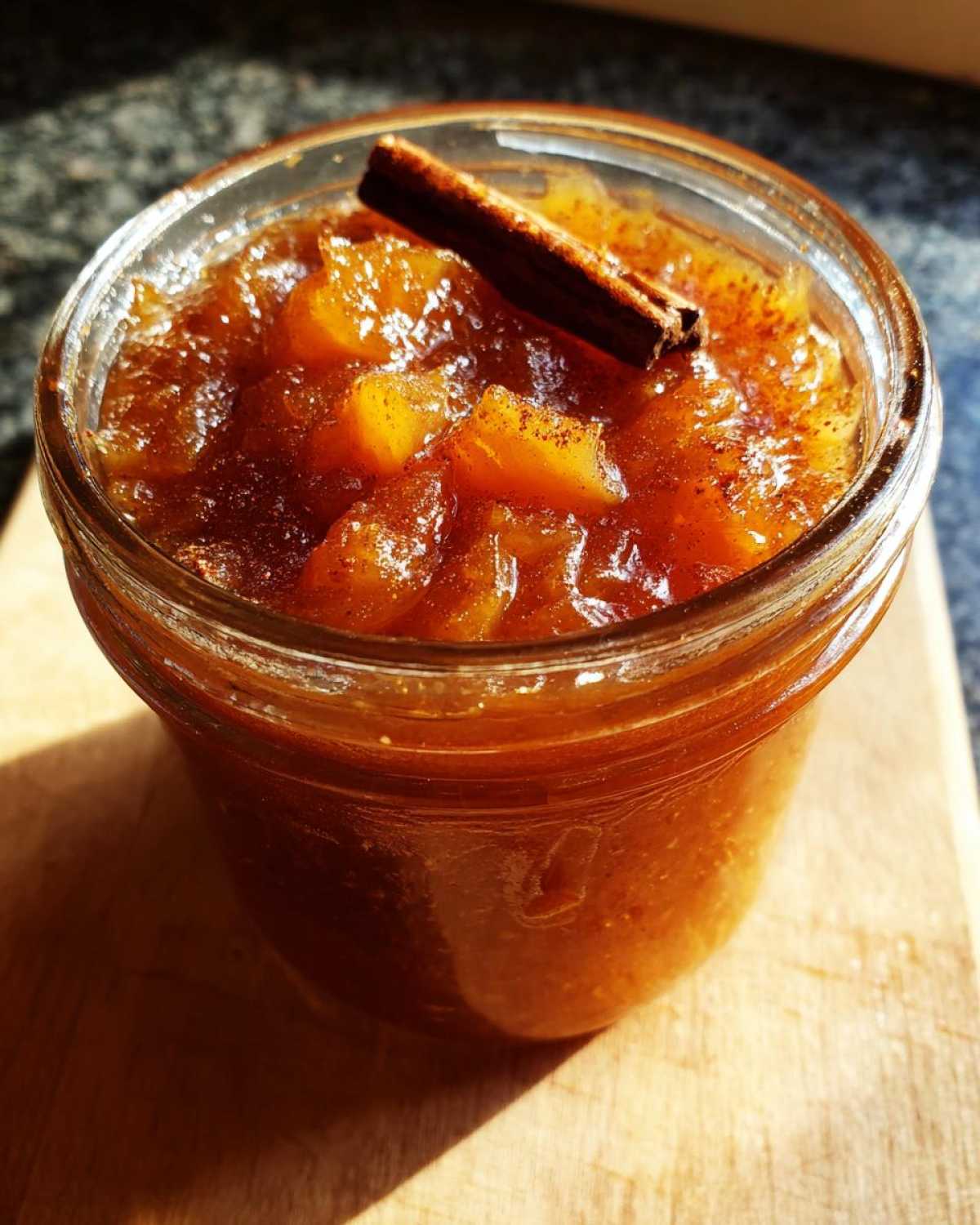 A jar of homemade pear preserves, garnished with a cinnamon stick, sits on a wooden board.