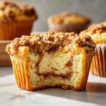 A close-up of a pumpkin cheesecake muffin with a bite taken out, showing the cheesecake swirl and crumb topping.