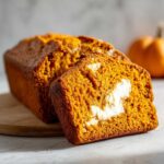 A sliced moist pumpkin loaf with a visible cream cheese filling, sitting on a wooden board.