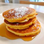 A stack of three pumpkin pancakes, topped with powdered sugar and drizzled with syrup.