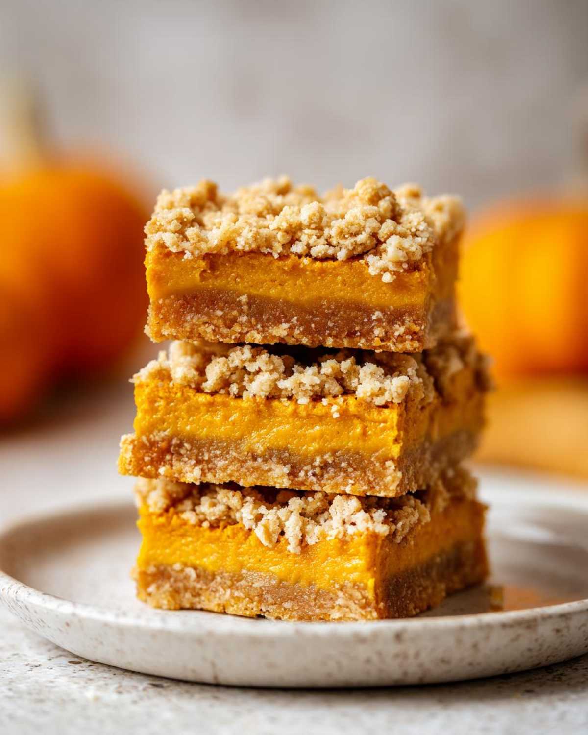 A stack of three pumpkin pie bars with a crumble topping, sitting on a speckled plate.