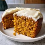 A slice of moist pumpkin spice cake with creamy white frosting on a white plate, ready to eat.