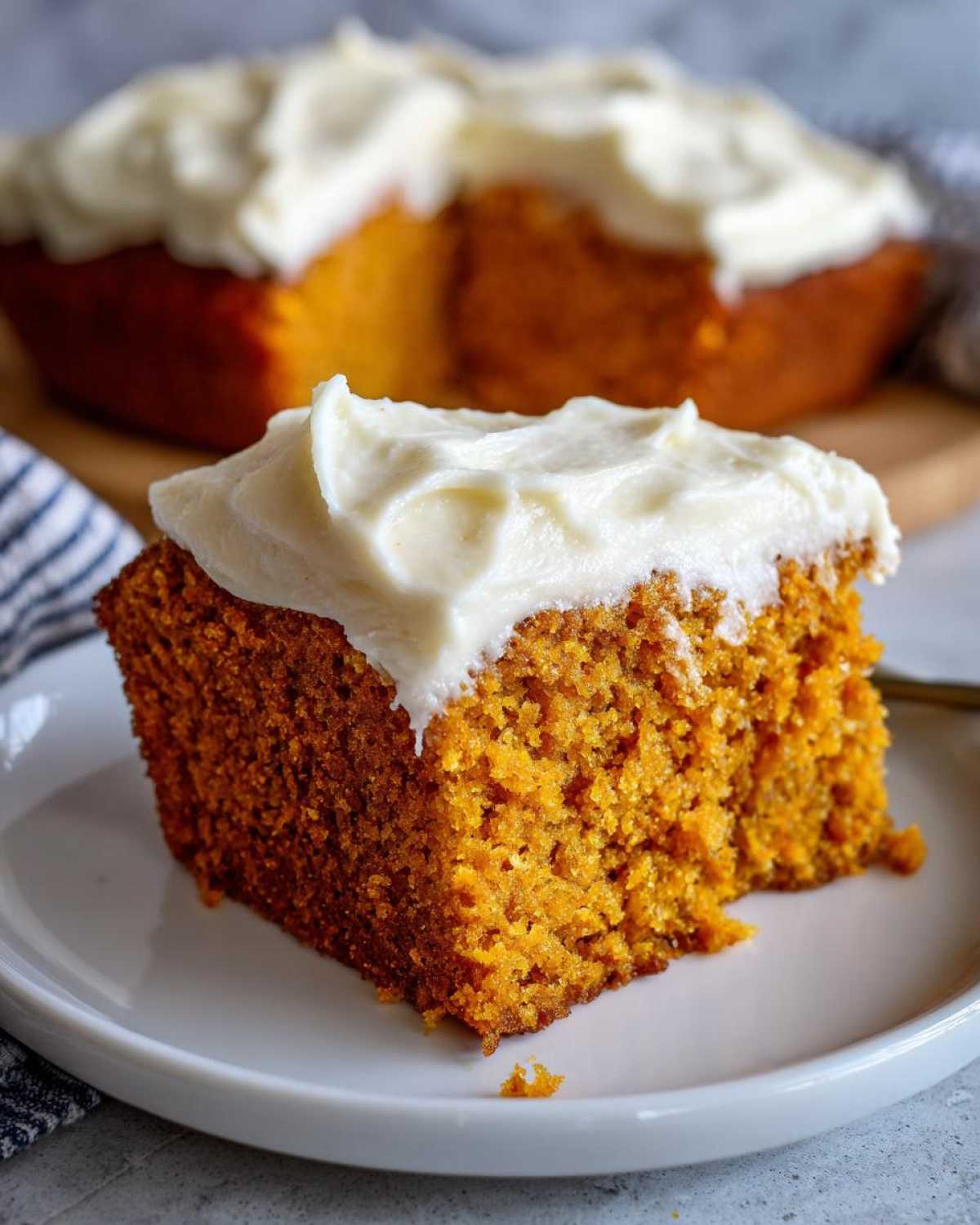 A slice of pumpkin spice cake topped with creamy white frosting on a white plate.