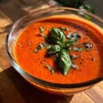 A bowl of vibrant tomato soup with fresh tomatoes, garnished with basil leaves and pepper.