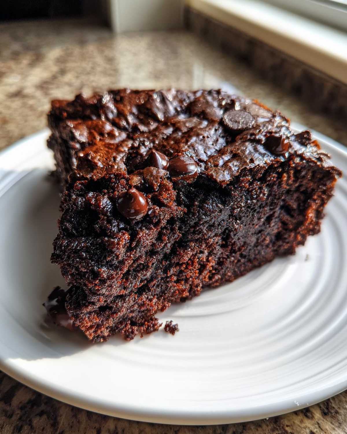 A single slice of fudgy zucchini brownies on a white plate, topped with chocolate chips.