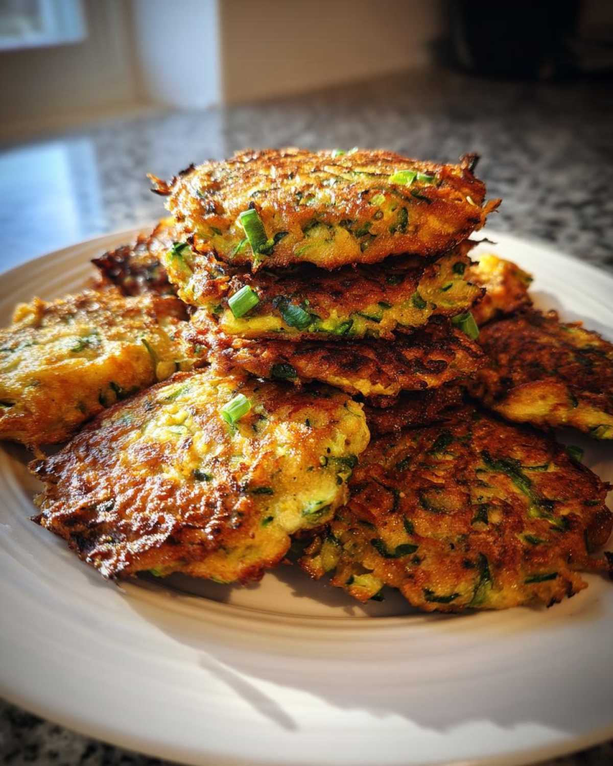 A stack of golden-brown zucchini fritters on a white plate, garnished with fresh green onions.
