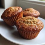Three freshly baked zucchini muffins arranged on a white plate, showcasing their golden-brown color and texture.