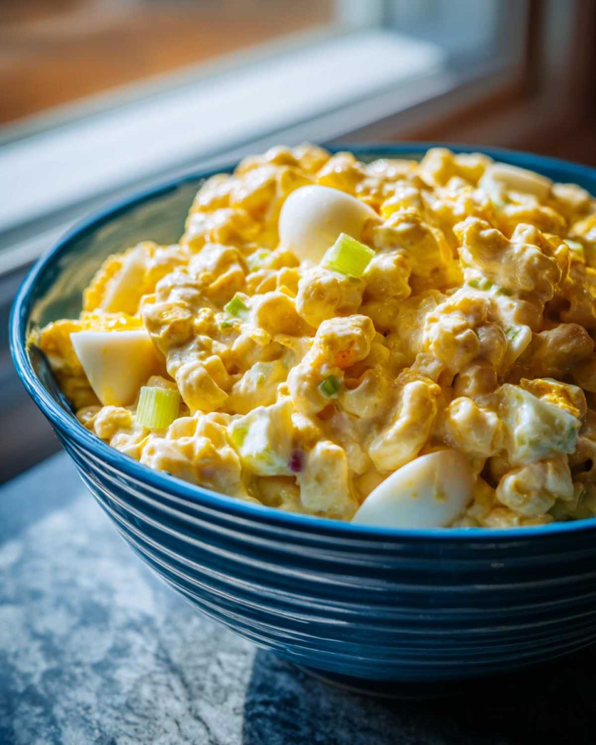 Close-up of Amish macaroni salad in a blue striped bowl, featuring macaroni, eggs, and creamy dressing.