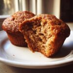 Two applesauce muffins on a white plate, one with a bite taken out showing the moist interior.