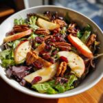 Close-up of a vibrant autumn salad with mixed greens, apple slices, pecans, and cranberries, drizzled with dressing.
