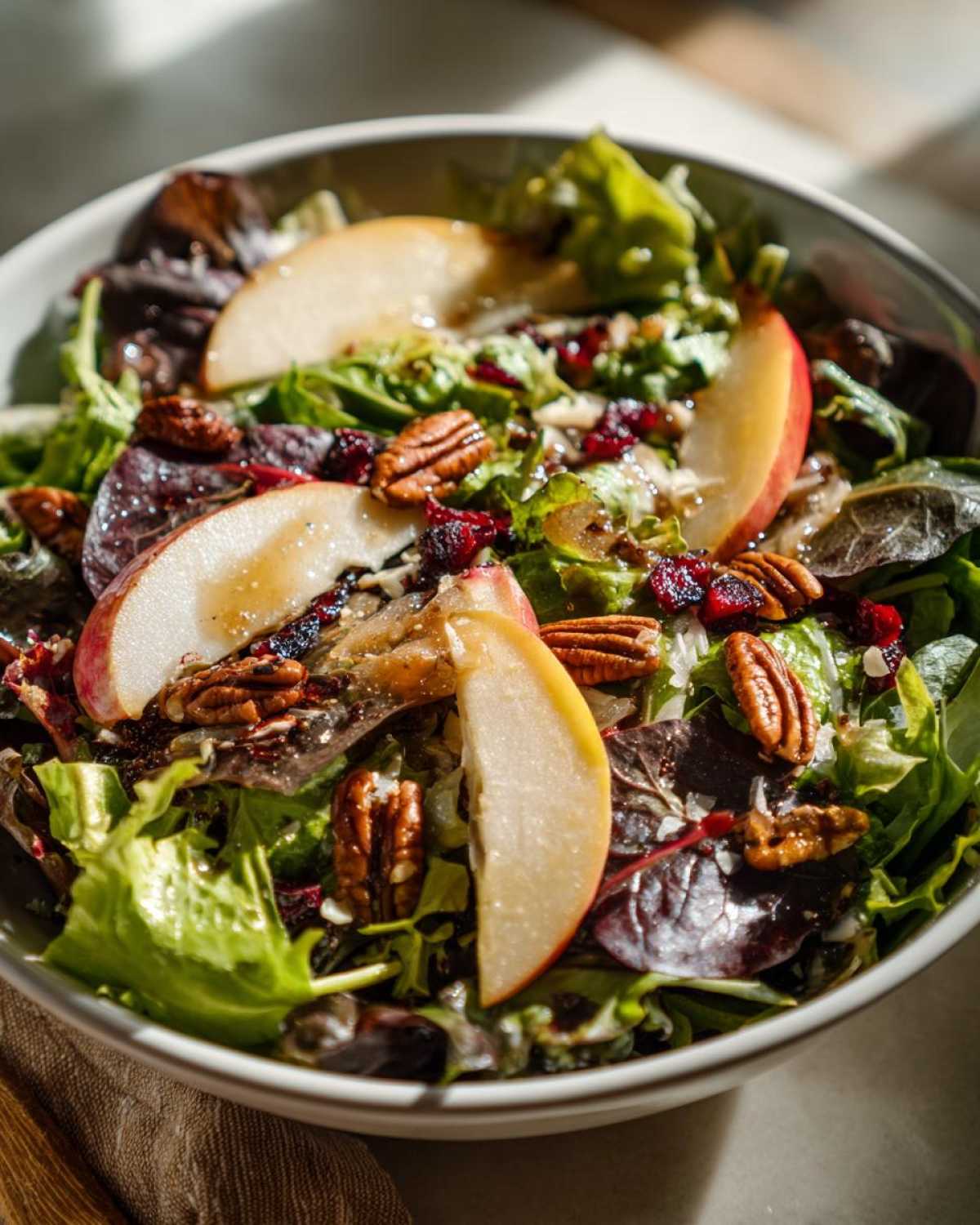 A vibrant autumn salad featuring mixed greens, apple slices, pecans, and cranberries in a white bowl.