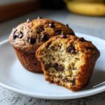 Two banana chocolate chip muffins on a white plate, one with a bite taken out to show the inside.