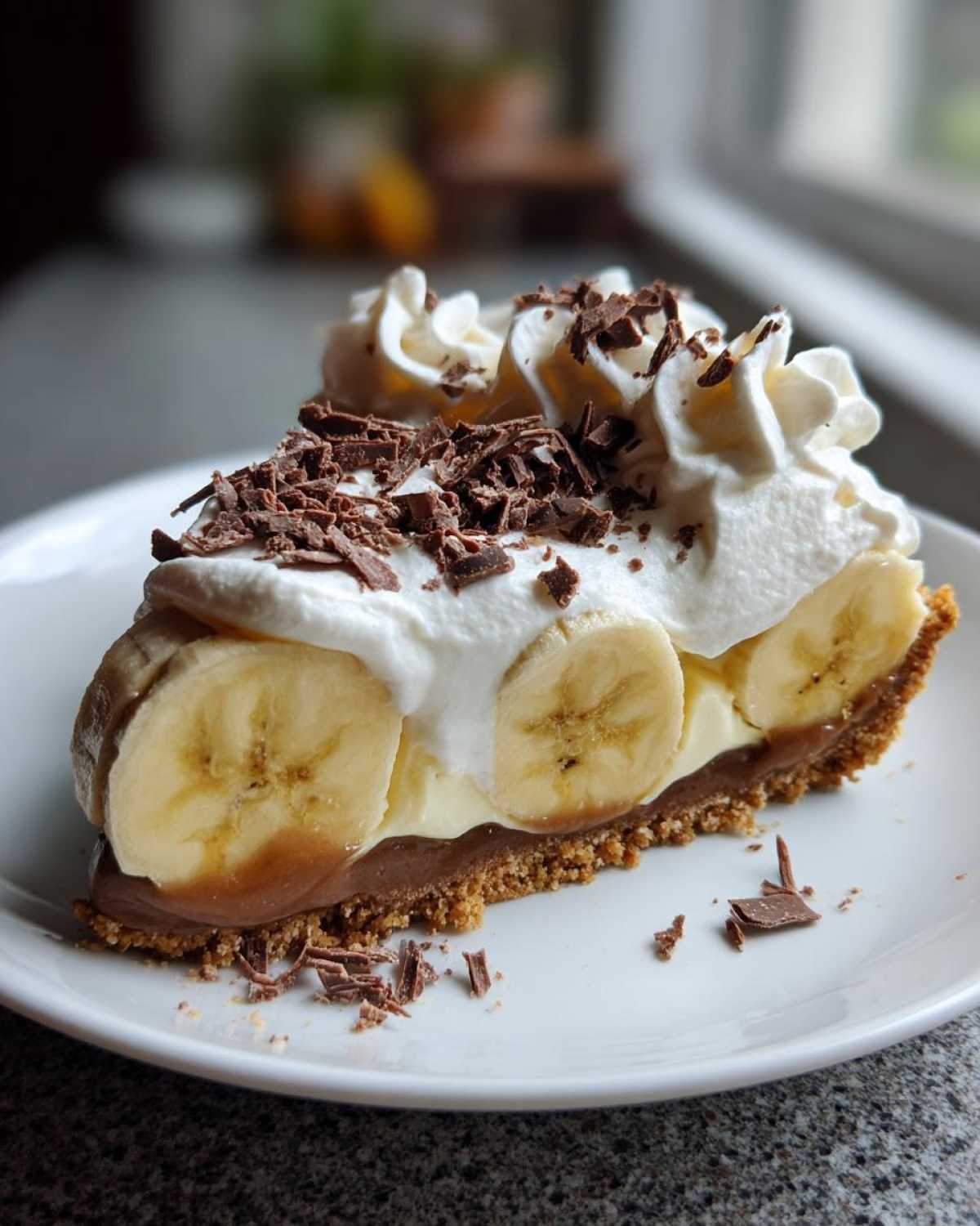 A slice of banoffee pie with bananas, whipped cream, chocolate shavings, and a graham cracker crust.