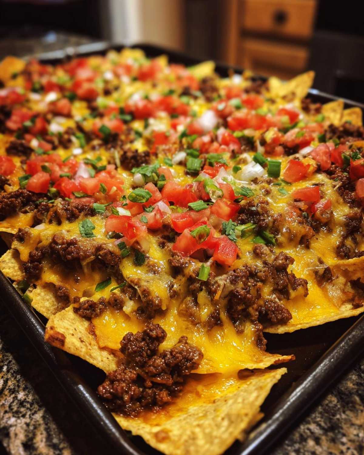 A baking sheet piled high with beef nachos, melted cheese, tomatoes, and green onions.