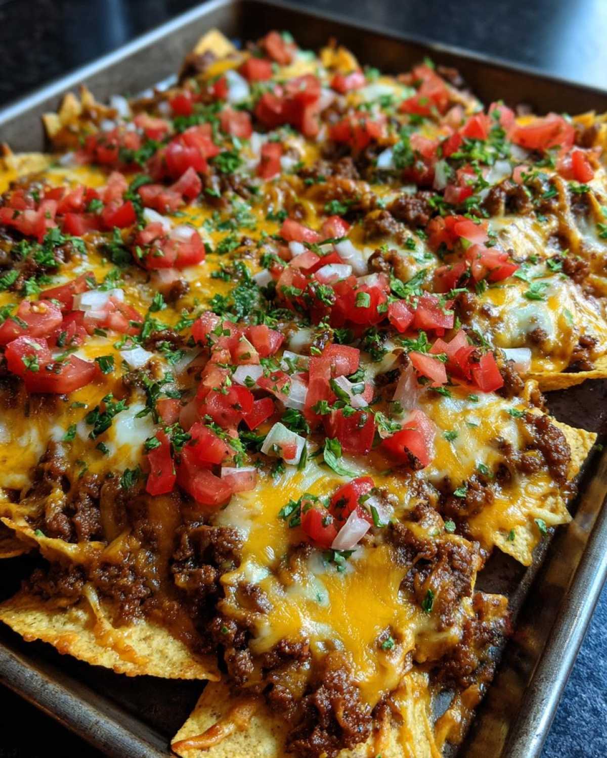 A baking sheet filled with beef nachos, melted cheese, diced tomatoes, onions, and parsley.