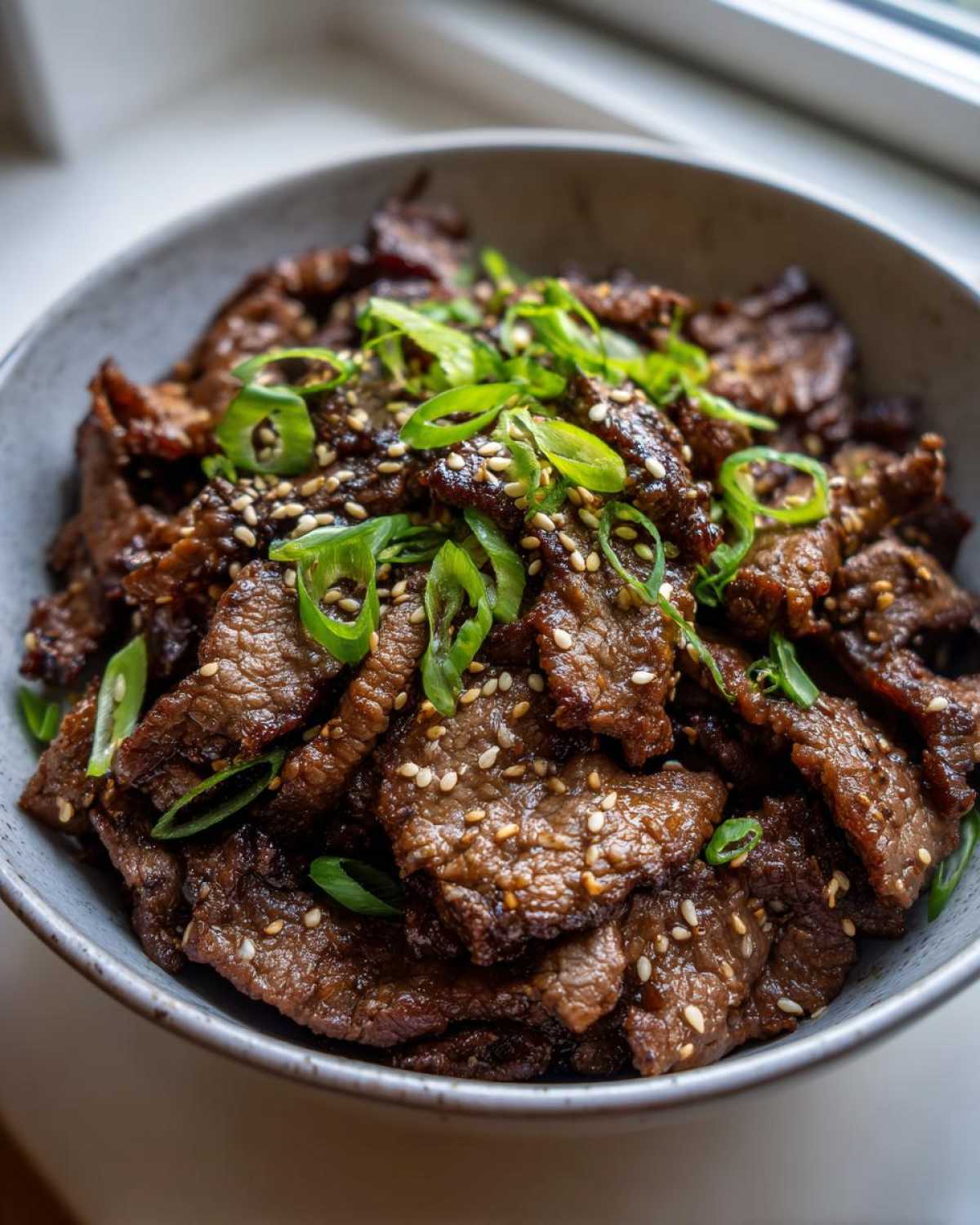 A bowl of Bulgogi recipe featuring thinly sliced, marinated beef, garnished with sesame seeds and green onions.