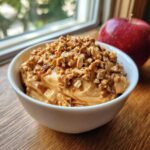 A bowl of caramel apple dip topped with chopped nuts, next to a red apple.
