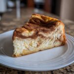 A delicious slice of cinnamon roll cheesecake on a white plate, showcasing the cinnamon swirl and creamy filling.