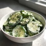 A white bowl filled with creamy cucumber salad, garnished with fresh dill.