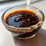 Close-up of flavorful dumpling sauce in a clear glass bowl, featuring sesame seeds and chili flakes.