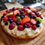 Delicious fruit pizza sugar cookie topped with whipped cream, strawberries, blueberries, raspberries, and kiwi.