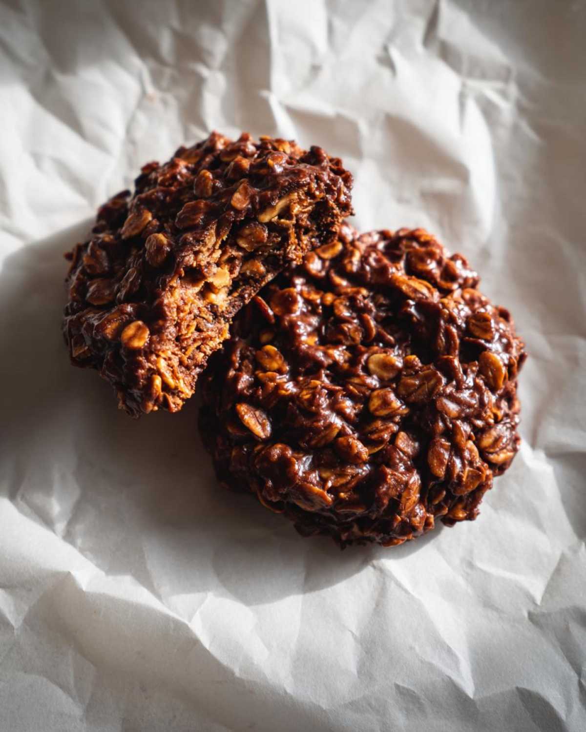 Two no-bake chocolate oatmeal cookies, one broken in half, on parchment paper.
