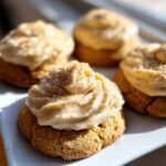 Four pumpkin cheesecake cookies with creamy frosting and spice dusting on a white plate.
