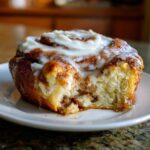 Close-up of a single pumpkin cinnamon roll with cream cheese frosting on a white plate, a bite taken out.
