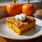 A slice of pumpkin dump cake on a plate, topped with whipped cream. Pumpkins are in the background.
