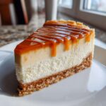 A slice of salted caramel cheesecake with a graham cracker crust and caramel drizzle on a white plate.