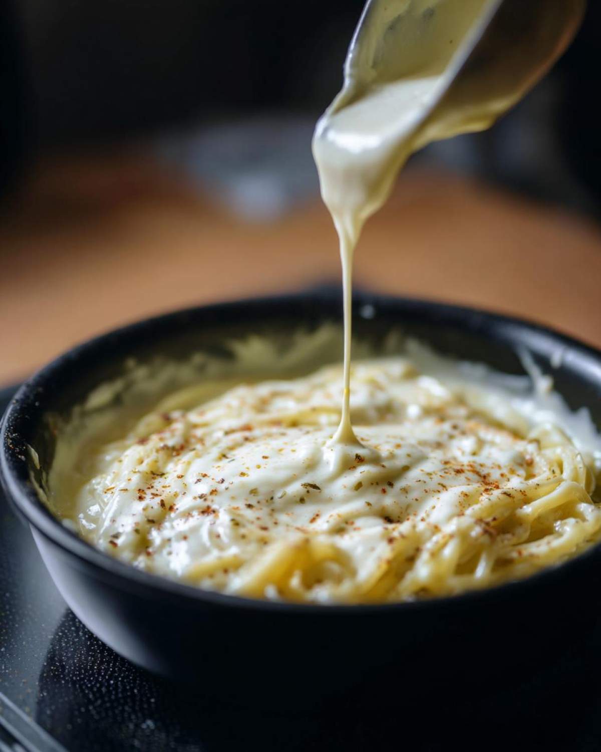 A spoonful of creamy alfredo sauce being poured over a bowl of pasta, seasoned with herbs and spices.