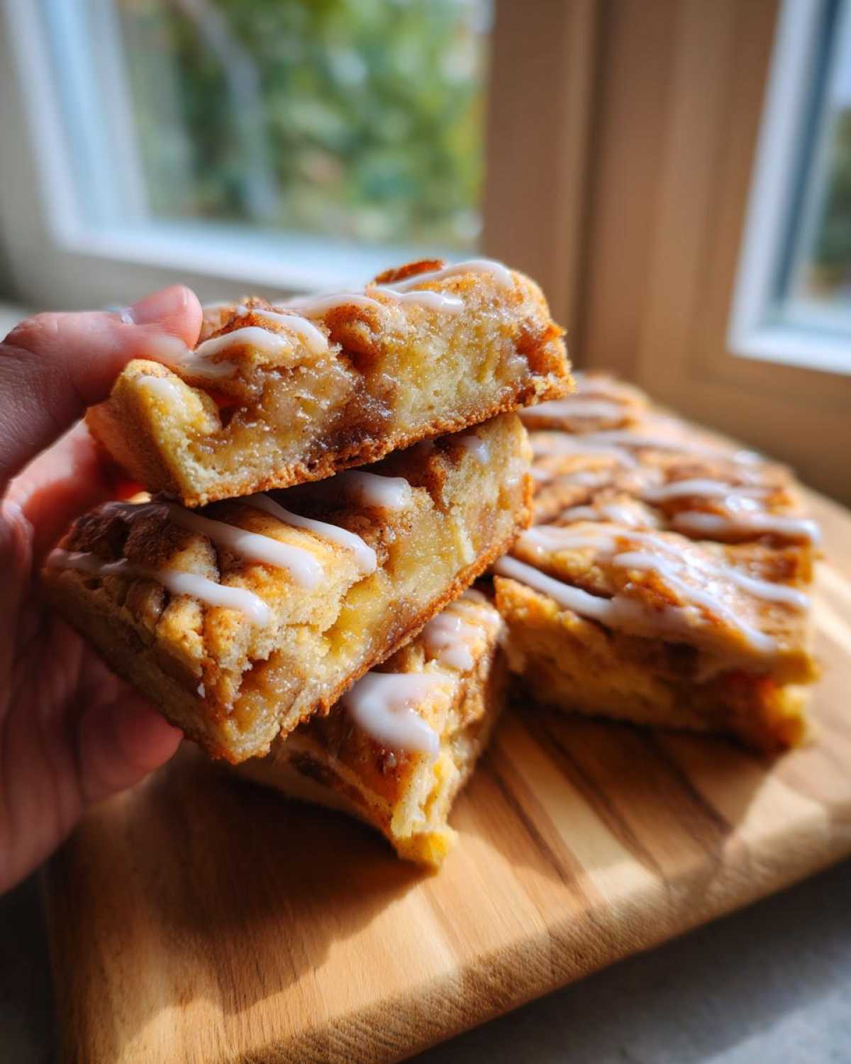 A hand holds a stack of three apple slice bars drizzled with icing, showcasing the layers of apple filling.