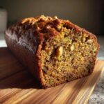 A loaf of homemade applesauce zucchini bread, studded with walnuts, sits on a wooden cutting board.