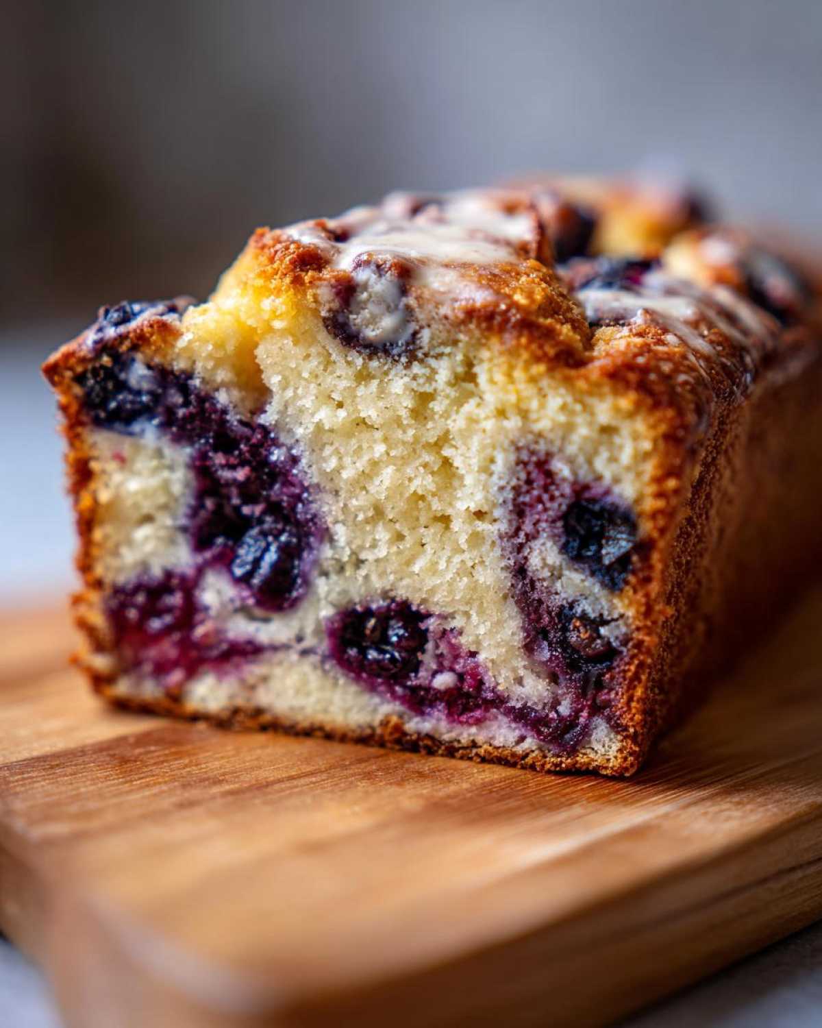 A close-up of a slice of blueberry cream cheese bread on a wooden board, showing blueberries and glaze.