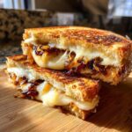 Stacked caramelized onion grilled cheese sandwich with melted cheese oozing out, on a wooden board.