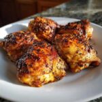 Four crispy baked chicken thighs arranged on a white plate, showcasing their golden-brown skin.
