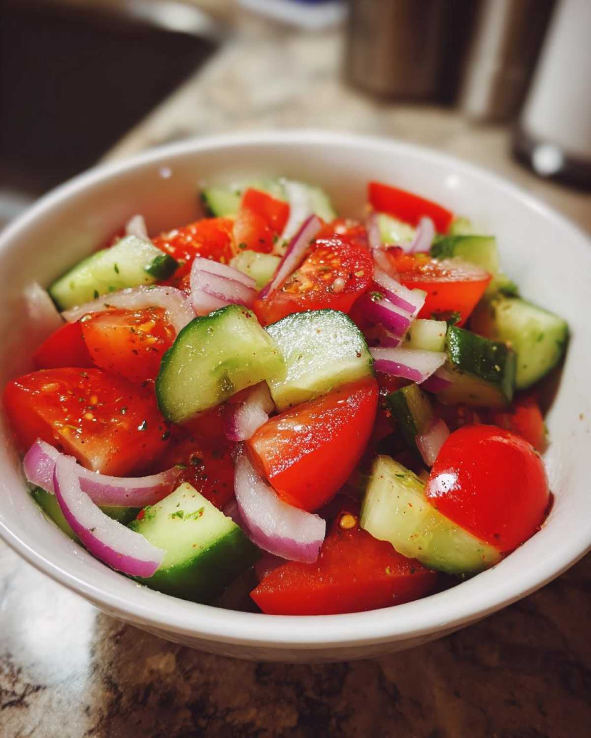 A vibrant cucumber tomato salad in a white bowl, featuring cucumbers, tomatoes, and red onions.