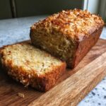 A loaf of Hawaiian banana bread with toasted coconut, sliced on a wooden board.