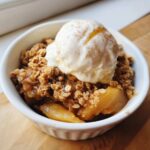 A white bowl filled with healthy apple crisp, topped with a scoop of vanilla ice cream and a sprinkle of cinnamon.