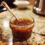 Close-up of homemade apple butter in a glass jar with a spoon, showcasing its rich color and texture.