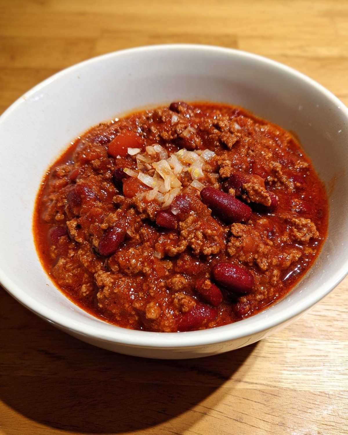 A bowl of hearty homemade chili, topped with diced onions, sits on a wooden surface.